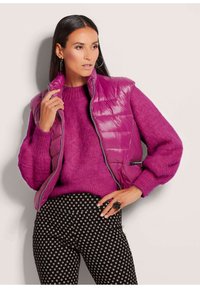 Magenta Steppweste mit horizontalem Quiltmuster, kombiniert mit einem flauschigen magentafarbenen Pullover und schwarzen Hosen mit geometrischem weißen Muster.