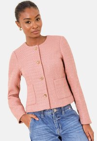 Veste texturée rose à manches longues, boutons dorés, col rond et deux poches avant, portée avec un jean bleu.