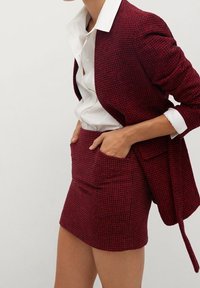 Blazer rosso con texture e gonna coordinata, con una camicia bianca senza colletto sotto. La gonna ha tasche frontali e un design aderente.