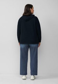 Frau, die mit dem Rücken zur Kamera steht, trägt einen schwarzen Hoodie, blaue Jeans und weiße Sneaker vor einem schlichten hellgrauen Hintergrund.