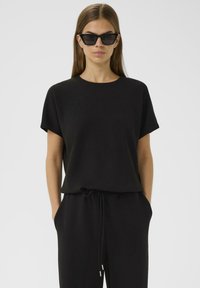 Schwarzes Kurzarmhemd mit einem runden Halsausschnitt, kombiniert mit schwarzen Jogginghosen mit Kordelzug. Das Model trägt eckige schwarze Sonnenbrillen. Glatte Stofftextur.