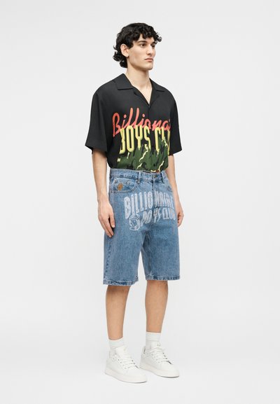 Fiatal férfi fekete, rövid ujjú ingben, piros és sárga felirattal, fehér nyomtatással díszített farmer rövidnadrágban, fehér zokniban és fehér tornacipőben.