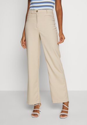 Kobieta ubrana w beżowe, szerokie spodnie z wysokim stanem, niebiesko-biały prążkowany top bez rękawów oraz białe sandały na obcasie z paskami, stojąca na szarej podłodze.