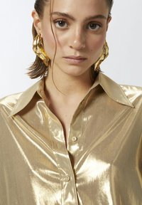 Zlatna metalik košulja dugih rukava sa glatkom, sjajnom teksturom i klasičnom ovratnikom. Velike zlatne skulpturalne naušnice imaju teksturirani dizajn.