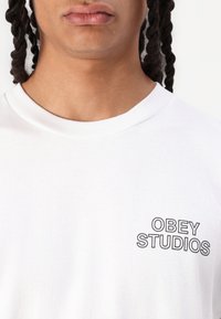 White cotton t-shirt with a round neckline, featuring black printed text "OBEY STUDIOS" on the left chest. Soft texture, casual design.