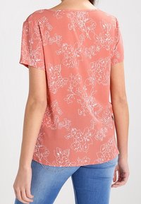 Blouse corail à motif floral blanc, manches courtes, encolure ronde, et tissu léger, associée à un jean en denim bleu.