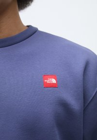 Sudadera púrpura con cuello redondo y puños acanalados. Presenta una pequeña etiqueta roja con el logotipo "THE NORTH FACE" en el área del pecho.