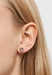 Primo piano di un orecchio sinistro con capelli biondi, che indossa un piercing industriale in oro e un orecchino a forma di cuore.