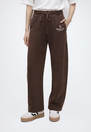 Persona che indossa pantaloni della tuta marroni con coulisse e scarpe da ginnastica bianche, in piedi con una mano in tasca su sfondo semplice.