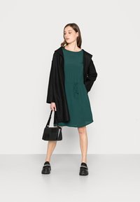 Vestido verde com cordão na cintura, sobreposto a um casaco preto. Bolsa preta e sapatos robustos completam o conjunto. Texturas lisas por toda a parte.