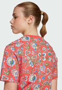 Korallfärgad bomulls-t-shirt med ett blommönster i blå och gröna nyanser, korta ärmar och rund halsringning. Tygets struktur verkar slät.