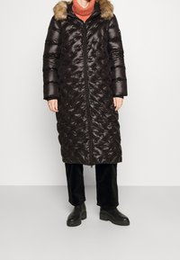 Abrigo largo acolchado negro con capucha con borde de piel, con cierre de cremallera, diseño acolchado y bolsillos laterales, combinado con pantalones de pana negros y botas.