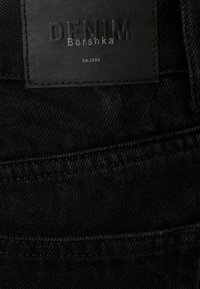 Tela de mezclilla negra con superficie texturizada. Presenta una etiqueta tejida con "DENIM" y "Bershka" en relieve, y detalles de costura.