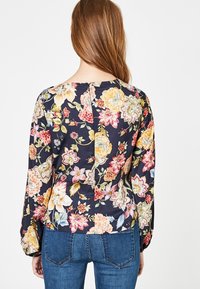 Blouse florale avec un fond noir, manches longues, fleurs colorées en rose, jaune et vert, avec un détail subtil d'ouverture dans le dos.