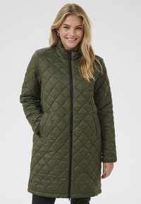Manteau matelassé, longueur genoux, de couleur vert olive. Il possède un col haut, une fermeture éclair à l'avant et des poches latérales avec une texture lisse et brillante.