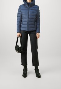 Giacca a vento imbottita blu navy con cappuccio, caratterizzata da un design trapuntato orizzontale, zip frontale, abbinata a jeans neri e stivali, con una borsa nera in mano.