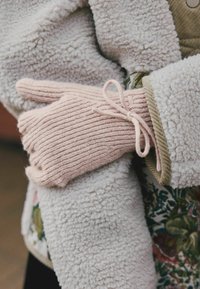Guanto rosa a maglia con texture a costine e un fiocco decorativo, indossato sopra un giubbotto in sherpa beige con fodera floreale. La mano destra è leggermente aperta.