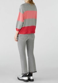 Femme portant un pull rayé gris et rouge, un jean gris cropped, des chaussettes noires et des mocassins argentés, se tenant devant un fond clair.