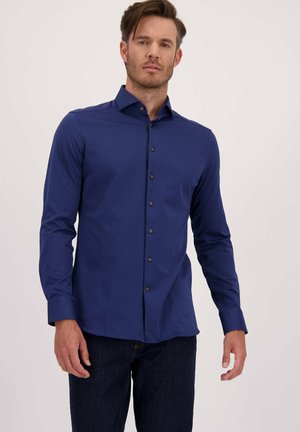 Man in een aansluitend, donkerblauw overhemd met lange mouwen en een donkere jeans, staand tegen een effen lichte achtergrond.