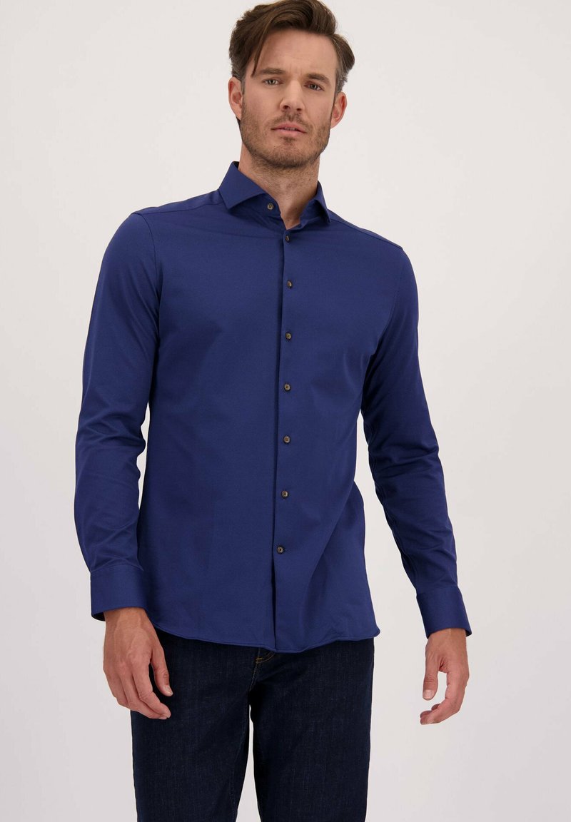 Man in een aansluitend, donkerblauw overhemd met lange mouwen en een donkere jeans, staand tegen een effen lichte achtergrond.