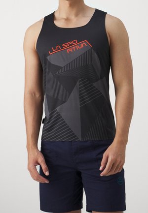 Mężczyzna ubrany w czarny top na ramiączkach z czerwonym napisem i geometrycznymi wzorami, w połączeniu z ciemnoniebieskimi szortami z małym logo na lewej nodze.