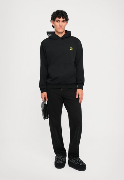 Haut à capuche noir avec un logo de smiley jaune, cordon de contraste, poche avant, associé à un pantalon noir et des chaussures au motif unique.