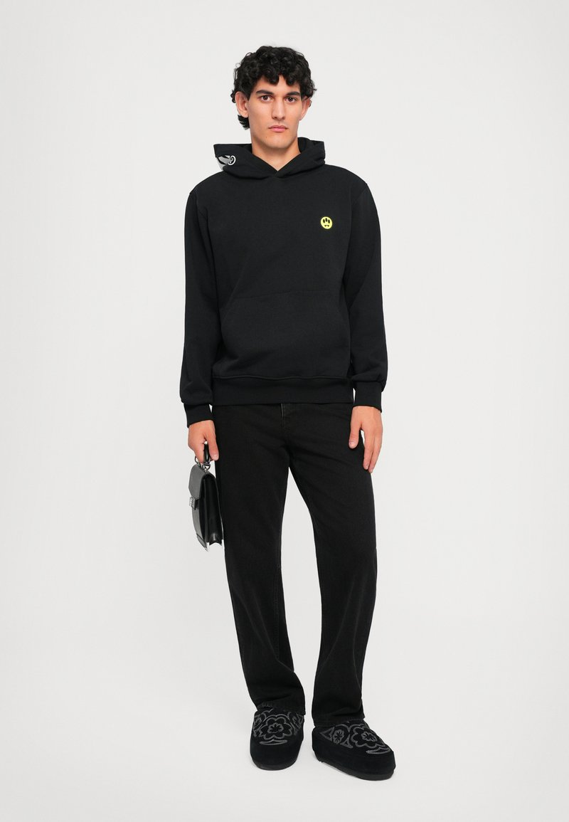 Felpa nera con logo di un viso sorridente giallo, cordini in contrasto, tasca frontale, abbinata a pantaloni neri e calzature con un motivo unico.