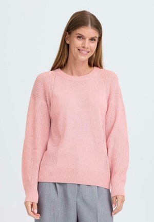 Ljusrosa stickad tröja med rund halsringning, raglanärmar och texturerade sömmar; kombinerad med grå byxor.