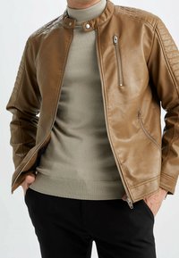 Uomo che indossa una giacca in pelle color beige con zip sopra un maglione leggero color beige, con le mani nelle tasche dei pantaloni neri, mostrato dalla spalla ai fianchi.