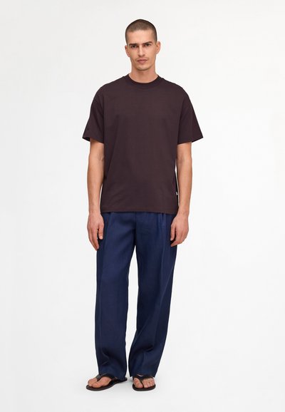 Homme debout portant un t-shirt marron foncé uni, un pantalon ample bleu marine et des tongs noires sur fond blanc.