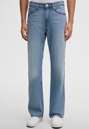 Mężczyzna ubrany w jasnoniebieskie jeansy o prostych nogawkach i białe buty wsuwane, stojący na tle jednolitego tła.