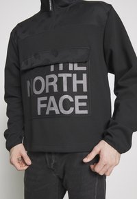 Pullover in fleece nero con colletto alto, tasca in rete e grande logo riflettente "THE NORTH FACE". Texture liscia e vestibilità rilassata.
