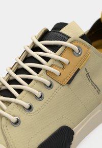 Μπεζ αθλητικό παπούτσι με υφασμάτινη υφή, μαύρες λεπτομέρειες και κρεμ κορδόνια. Διαθέτει κίτρινη δερμάτινη ετικέτα και ασημένιες τρύπες για τα κορδόνια. Λαστιχένια σόλα με αυλακώσεις.