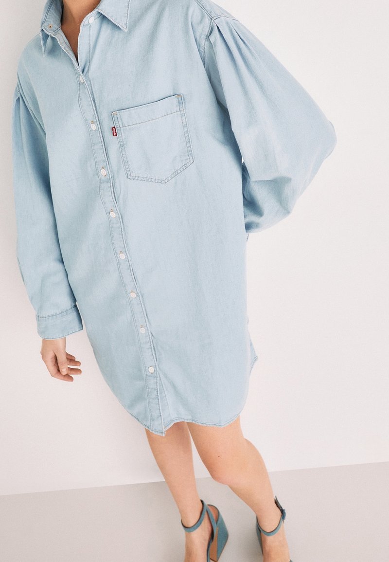 Chemise en denim bleu clair avec fermeture à boutons, manches larges et poche poitrine ; présente un ourlet arrondi et une texture douce.