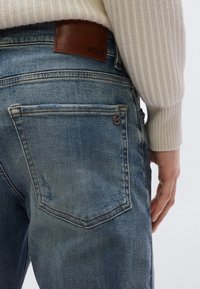 Koptatott kék farmernadrág barna bőr BOSS címkével hátul. Két hátsó zsebe van, minimális kopás részletekkel.