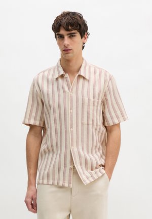 Mann trägt cremefarbenes kurzärmeliges Hemd mit vertikalen roten Streifen und beigefarbene Hose, steht vor weißem Hintergrund.