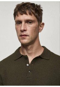 Dark olive green knit polo shirt with a collar, three-button placket, and ribbed texture, worn by a model with short hair.