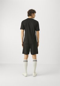 T-shirt sport negru, pantaloni scurți negri, șosete albe cu dungi negre și ghete albe. Văzut din spate, evidențiind un design ajustat.