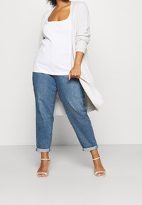 Dlouhý světle šedý kardigan přes bílý tílko, v kombinaci s modrými džínami s ohrnutými nohavicemi a tělovými sandály na podpatku. Měkká pletená textura.