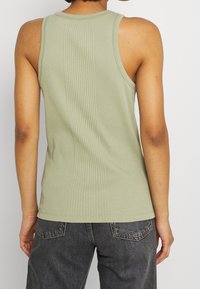 Oliwkowo-zielony prążkowany top na ramiączkach o dopasowanym kroju, z grubymi ramiączkami i pionowymi przeszyciami z tyłu. Noszony z ciemnymi dżinsami.