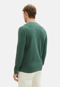 Dunkelgrüner Strickpullover mit langen Ärmeln, rundem Ausschnitt und gerippten Bündchen sowie Saum. Die Textur wirkt weich und warm.