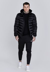 Chaqueta acolchada negra con cremallera frontal y capucha, combinada con pantalones de chándal negros ajustados con bolsillos laterales y zapatillas blancas.