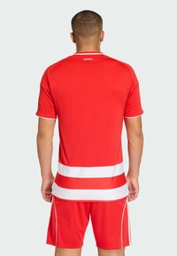 Červený športový dres s bielymi detailmi a vodorovnými pruhmi, krátkymi rukávmi a okrúhlym výstrihom. Nosí sa so zodpovedajúcimi červenými šortkami.