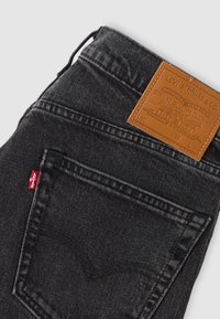 Μαύρο τζιν από denim με δερμάτινο εμπορικό σήμα της Levi's και κόκκινη ετικέτα ύφασμα της Levi's στην πίσω τσέπη, πάνω σε φωτεινό φόντο.