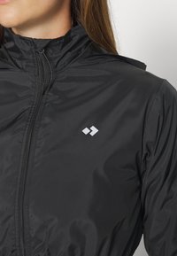 Sort letvægtsjakke med høj krave, lynlås fortil og et lille hvidt logo. Har en glat, blank tekstur og en tætsiddende pasform.