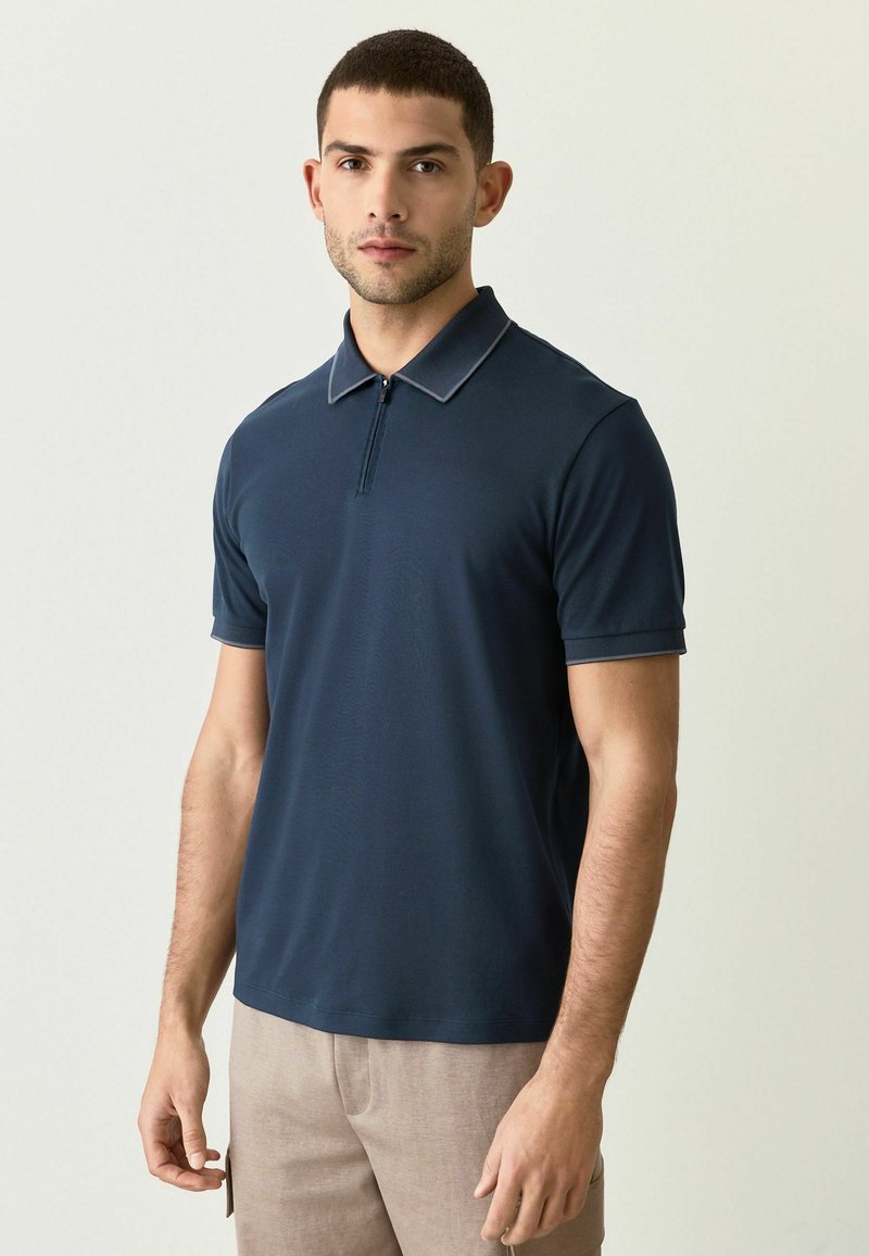 Un hombre está de pie en una camiseta polo ajustada de color azul marino con cuello de cremallera, con las manos relajadas a los lados, frente a un fondo suave y neutro.