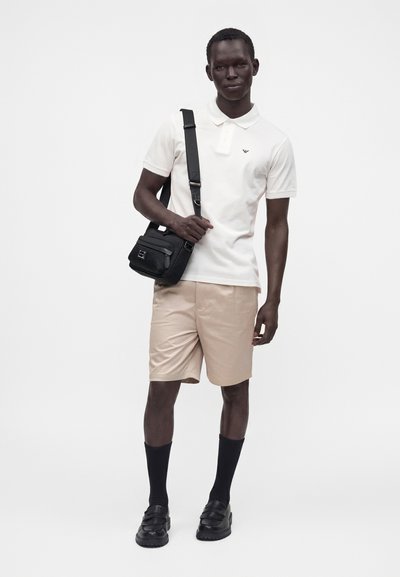 Homme portant un polo blanc, un short beige, des chaussettes noires, des mocassins noirs et un sac bandoulière sur fond blanc uni.
