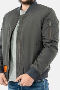 Bomberjack in donkergrijs nylon, voorzien van ribbelmanchetten, voorrits, zijzakken en een oranje label aan de linkerkant.