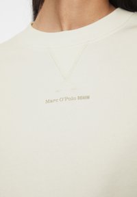 Sudadera de color crema con un cuello redondo texturizado, que presenta un sutil bordado y un pequeño logo marrón claro en la parte delantera.