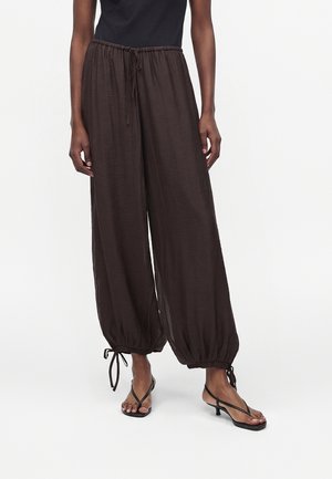 Osoba nosiaca čierne voľné nohavice so šnúrkou a zbieranými členkami a čierne remienkové sandále s nízkym opätkom na jednoduchom pozadí.